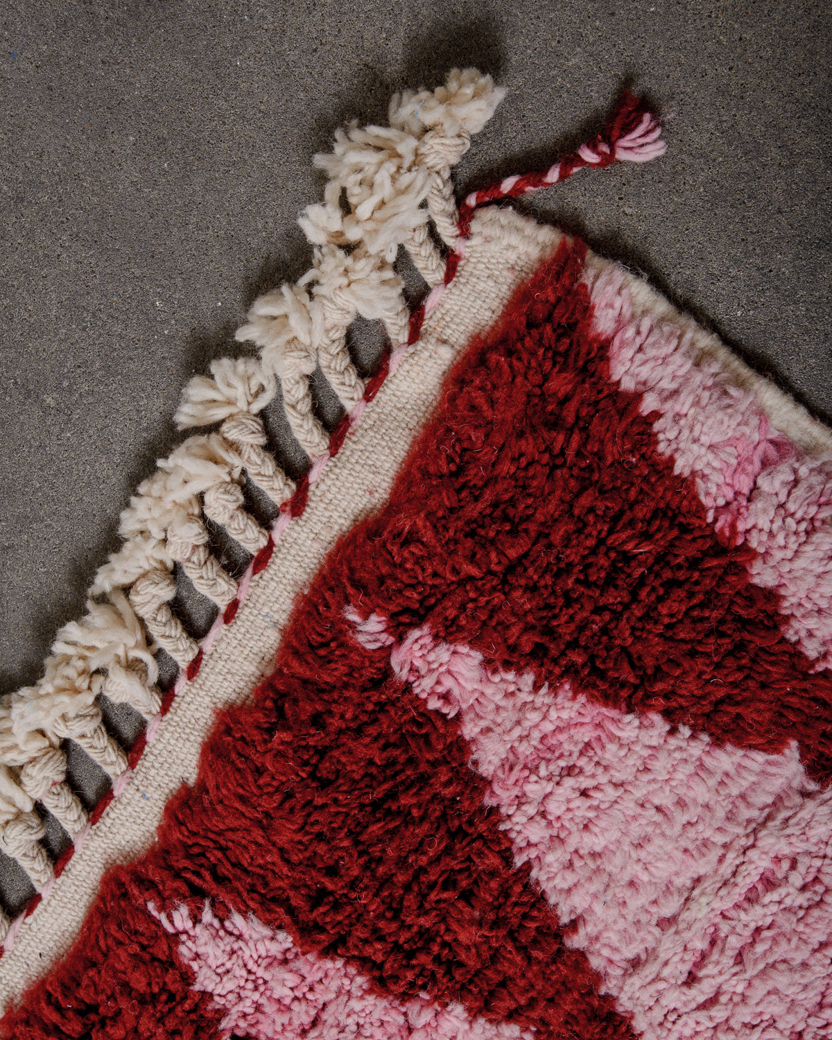 Corner of fluffy Berberlin berber rug in pink and burgundy wool and with natural wool tassels, laying on concrete floor.